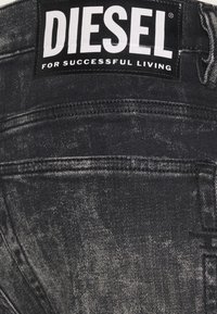 Jeans de mezclilla negros con una textura desgastada y una etiqueta negra prominente con el texto blanco "DIESEL PARA UNA VIDA EXITOSA."