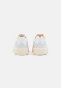 Baskets en cuir blanc avec des accents jaunes et un dos texturé. Elles disposent d'une semelle épaisse crème et d'éléments de design ondulés le long du talon.