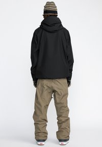 Svart vattentät jacka med huva, tillsammans med bruna snöbyxor med förstärkta knäpartier, samt en grå och beige randig mössa.