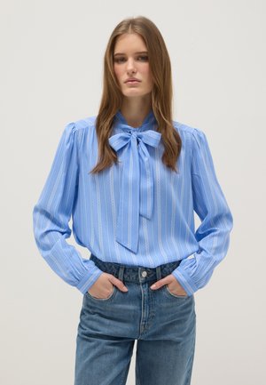 Jonge vrouw die een blauw gestreepte blouse met een striknek en een hoge taille blauwe spijkerbroek draagt, handen in de zakken, neutrale achtergrond.