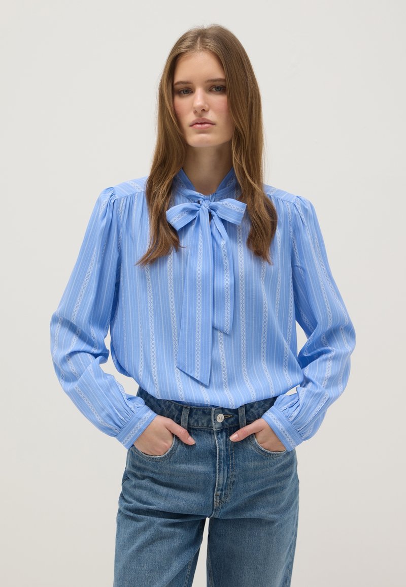 Young woman wearing a blue striped blouse with a bow tie collar and high-waisted blue jeans, hands in pockets, neutral background.