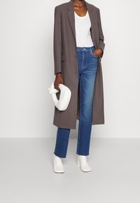 Blazer largo gris sobre una camiseta blanca, combinado con jeans de tiro alto azul y botas blancas hasta el tobillo. Sosteniendo un bolso acolchado blanco.