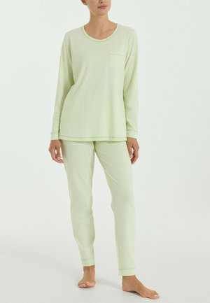 Donna che indossa una maglia a maniche lunghe verde chiaro e pantaloni abbinati, in piedi a piedi nudi contro uno sfondo bianco semplice.