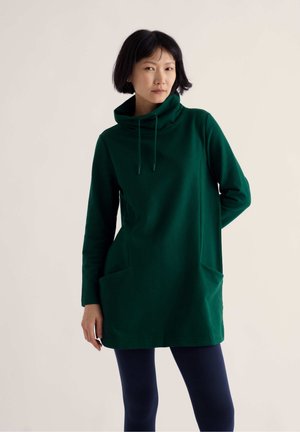 Femme portant une tunique à manches longues vert foncé avec un col bénitier haut et des poches, associée à des leggings foncés sur un fond clair.