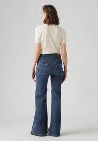 Light beige short-sleeve sweater paired with dark blue flared jeans featuring two back pockets and a leather patch on the waistband.