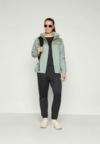 Kvinne iført grønn og svart utendørsjakke, svarte bukser, hvite sneakers, solbriller og bærende en svart ryggsekk mot en ensfarget bakgrunn.