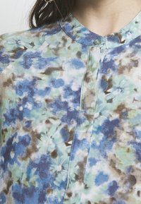 Gros plan d'une personne portant un chemisier avec un motif floral bleu, vert et marron et un petit col rond boutonné au niveau du cou.