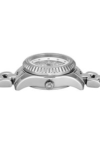 Montre métallique avec un boîtier rond en acier inoxydable, une lunette texturée et une couronne gravée, dotée d'un cadran transparent mettant en valeur des détails complexes.