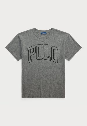 Pelēka kokvilnas T-kreklā ar īsām piedurknēm un apaļu kakla izgriezumu, ar vārdu "POLO" treknās melnās burtiem krūšu daļā.