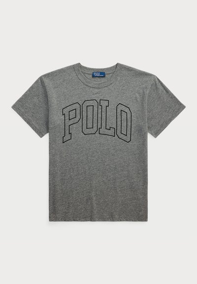 Pelēka kokvilnas T-kreklā ar īsām piedurknēm un apaļu kakla izgriezumu, ar vārdu "POLO" treknās melnās burtiem krūšu daļā.