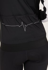 Schwarzes Sportoberteil mit einem weißen Herzschlag-Motiv, aus strukturiertem Stoff, enganliegendem Schnitt und gerafftem Saum hinten. Passende schwarze Leggings.