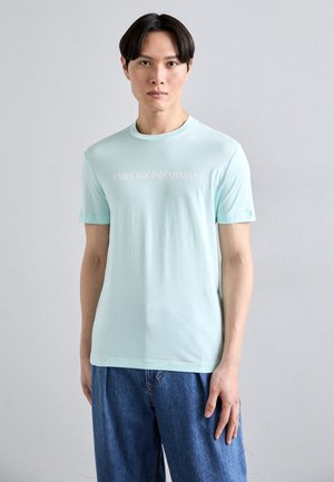 Camiseta de algodón de cuello redondo en azul claro que presenta un logo blanco de Emporio Armani en el pecho, combinada con unos pantalones vaqueros de corte holgado.