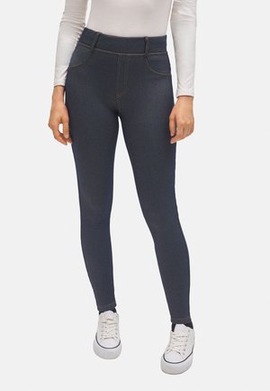Jeggings azul oscuro con una textura suave, cintura alta y bolsillos frontales falsos. Acentuados con costuras naranjas y combinados con zapatillas blancas.