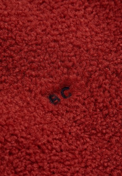 Tessuto in pile rosso con una superficie testurizzata e soffice. Presenta un piccolo ricamo blu con le lettere "BC" in basso a sinistra.