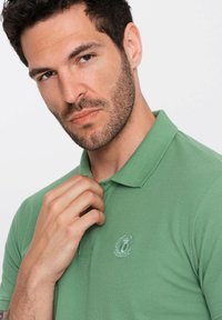 Zelená polo košile vyrobená z texturovaného materiálu, s ohrnovacím límcem a vyšívaným logem na levé hrudi. Model si upravuje límec.