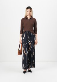 Polo shirt marron à manches longues cannelées associé à un pantalon large bleu marine avec un motif abstrait crème et doré. Chaussures noires à bout pointu et un sac marron.