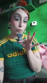 Grön Adidas-tröja med gula detaljer, håller i en liten svart mikrofon. Långa, färgglada akrylnaglar och en grön mönstrad huvudduk.