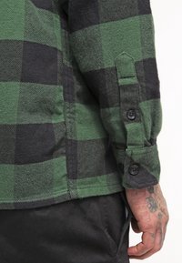 Grön och svart rutigt flanellskjorta med knappmanschett och sidosöm. Tyget verkar mjukt med en texturerad finish.