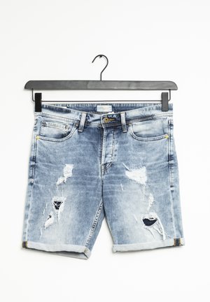 Shorts en jean bleu clair avec ourlets roulés et patches usés, suspendus à un cintre noir sur un fond blanc.