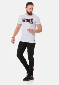 Weißes T-Shirt mit kurzen Ärmeln und fettem schwarzen "WORK"-Aufdruck; kombiniert mit schwarzen Slim-Fit-Hosen und schwarzen Sportschuhen.