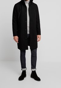 Homme portant un long manteau noir à boutons, un pull à col roulé gris, un jean foncé retroussé et des bottes en suède noires, se tenant devant un fond uni.