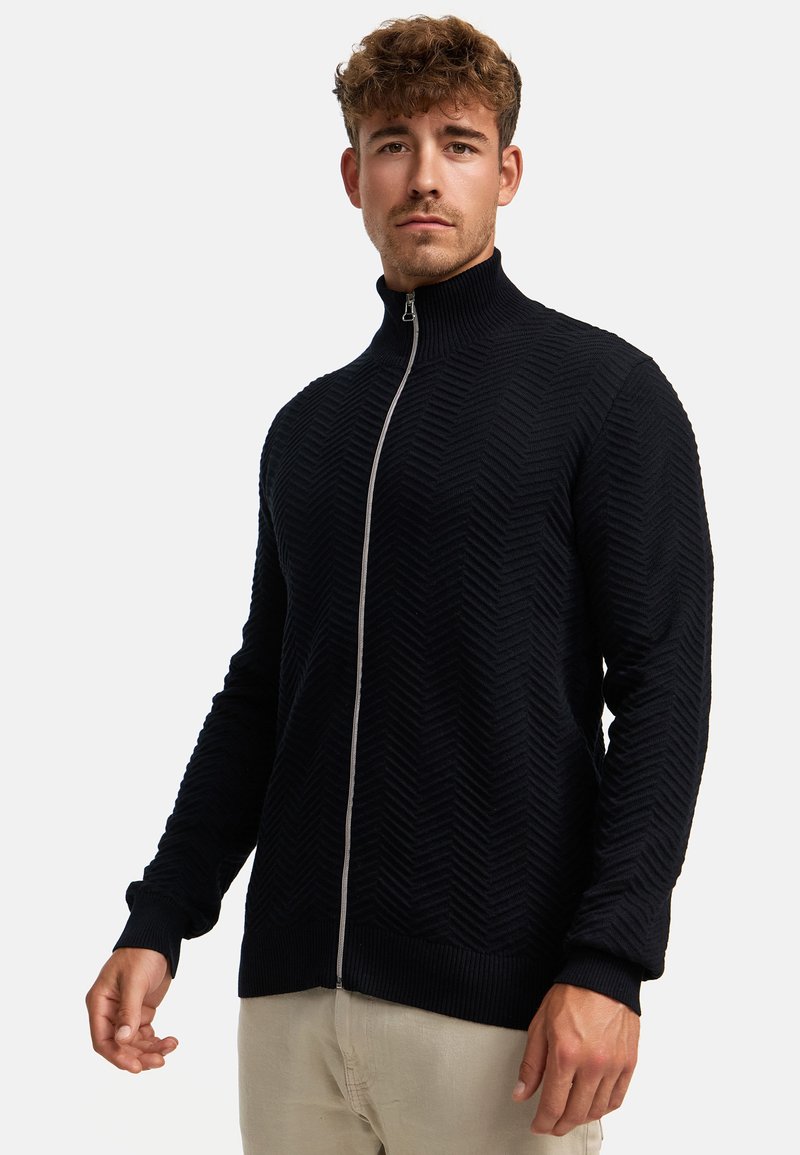 Sweater negro con cremallera fabricado en tejido texturizado que presenta un patrón de chevron. Tiene un cuello alto y puños y dobladillo acanalados.