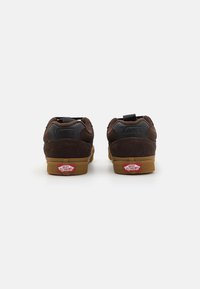 Baskets en daim marron avec une semelle extérieure en caoutchouc gomme, dotées d'accents noirs et d'une étiquette logo rouge Vans sur le talon. Design classique basse.