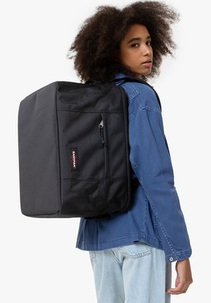 Sac à dos rectangulaire noir en tissu texturé, avec une poche avant et une fermeture éclair, porté sur une veste en denim bleue.