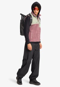 Fleece-Weste in Pink und Grün, darunter ein schwarzer Hoodie, schwarze Hose und ein schwarzer Rucksack. Das Modell trägt reflektierende Sonnenbrillen.