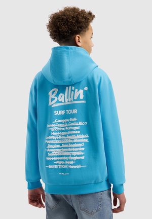 Jongere met krullend haar die een blauwe hoodie draagt waarop afgevinkte bestemmingen van de wereldwijde surftour op de achterkant staan.
