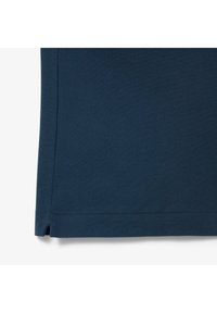 Tissu bleu foncé, gros plan sur l'ourlet d’un vêtement avec bord cousu et petite fente latérale sur fond blanc.