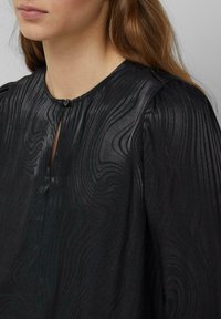 Kobieta ubrana w czarny top z delikatną falistą fakturą w paski i małym zapięciem na guzik przy dekolcie, widoczne długie brązowe włosy.