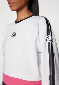 Sudadera gris con rayas horizontales negras y rosas. Presenta un cuello de canalé negro y un logo bordado en el pecho y la manga.