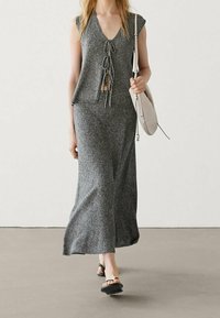 Frau in einem ärmellosen grauen Strickkleid mit Frontbindedetails, die eine beige Schultertasche trägt und schwarze Sandalen trägt, steht auf einem grauen Boden.