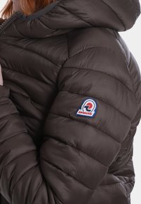 Giacca a vento marrone con cappuccio, caratterizzata da un design trapuntato, una texture liscia e un logo a patch sulla manica sinistra.
