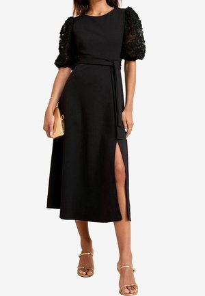 Robe midi noire avec manches bouffantes en dentelle florale et fente latérale, dotée d'une ceinture à la taille et d'une texture de tissu lisse. Accessoirisée avec une pochette dorée.