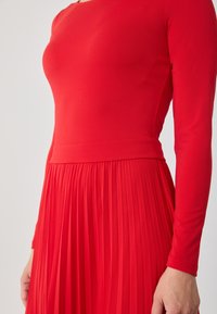 Rode lange mouwen jurk met een aansluitende bodice en geplooide rok. Gemaakt van een gladde stof, het heeft een ronde halslijn en een gedefinieerde taille.