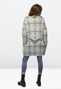 Cardigan gris de cuadros con mangas largas, que presenta un dobladillo redondeado y un detalle de bolsillo en la parte posterior. Combinado con jeans ajustados y botas negras de tobillo.