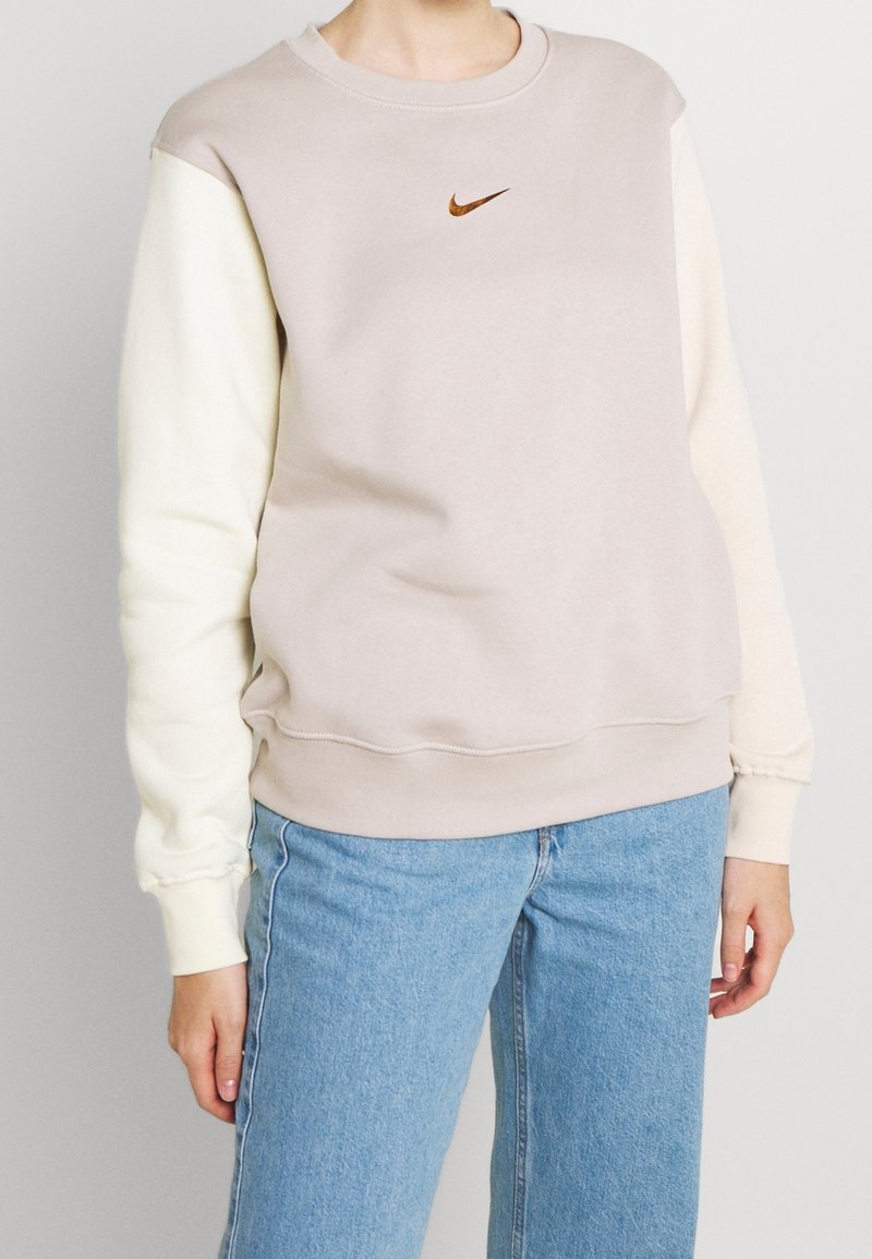 Nike-tröja med en beige kropp och krämfärgade ärmar. Har en liten brun logotyp på bröstet, ribbade manschetter och en rundad halsringning.