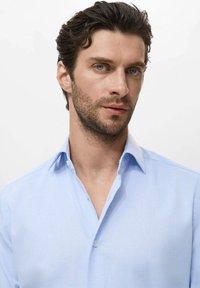 Man met kort donker haar en een lichte baard, gekleed in een lichtblauwe button-up shirt, die iets naar rechts kijkt op een witte achtergrond.