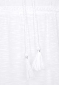 White cotton garment featuring braided drawstrings with metal accents, and white tassels at the ends. Textured fabric with subtle stripes.
