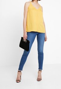 Top amarillo sin mangas con escote en V, combinado con vaqueros ajustados azules. La modelo sostiene un bolso negro con un asa dorada y lleva sandalias de tacón negras.