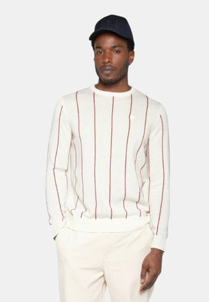 Pull crème avec des rayures verticales rouges, texture côtelée, col rond et manches longues. Associé à un pantalon clair et une casquette marine.