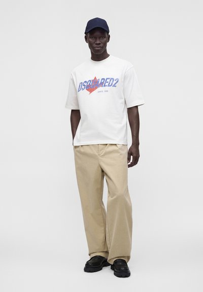 Άνδρας που φοράει λευκό t-shirt Dsquared2, μπεζ παντελόνι, μαύρα παπούτσια και σκούφο navy blue στέκεται μπροστά σε απλό φόντο.