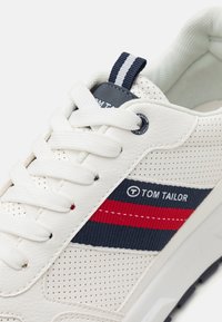 Baskets blanches avec un matériau synthétique perforé, des accents rayés bleu marine et rouge, et une étiquette de marque avec une boucle de traction au niveau du talon.