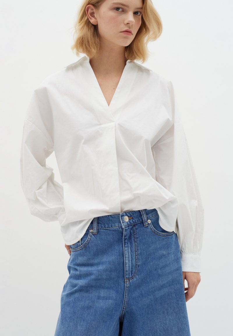 White cotton shirt with a deep V-neck and long, voluminous sleeves, paired with high-waisted blue denim shorts featuring a classic five-pocket design.