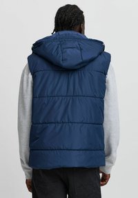 Tumši zilo mīksto bezmugurviedogu ar kapuci, kurai ir horizontāli izšūtas daļas un augsts apkakle, kārtota virs pelēka garām piedurknēm krekla.