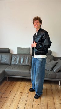 Μαύρο δερμάτινο μπουφάν τύπου bomber με ριμπ μανσέτες και φερμουάρ μπροστά, συνδυασμένο με φαρδιά μπλε τζιν και μαύρα παπούτσια, μπροστά σε ανοιχτόχρωμο τοίχο.