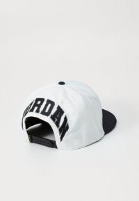 Casquette snapback blanche avec une visière noire et un logo "JORDAN" brodé à l'arrière. Fabriquée en tissu avec une texture lisse et une sangle réglable.