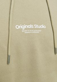 Oliwkowy hoodie z białym napisem "Originals Studio" oraz szczegółami brandingowymi. Posiada sznurki z metalowymi końcówkami i miękką teksturę.
